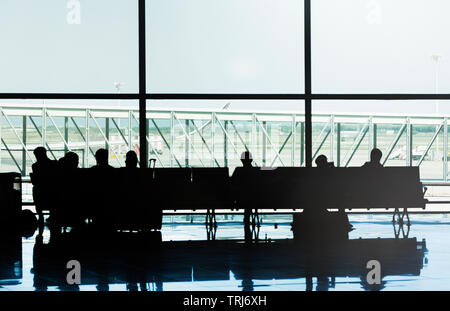 silhouettes of people sitting on the chairs of an airport waiting for their flight in front of the main window of the departure lounge Stock Photo