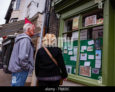 North Walsham, Norfolk, UK. 18 May 2019. Candid street photos – An older couple, a man in a grey hoodie and blue jeans and a lady in a black coat, reading the local classifieds in the window of a small shop in North Walsham high street Stock Photo