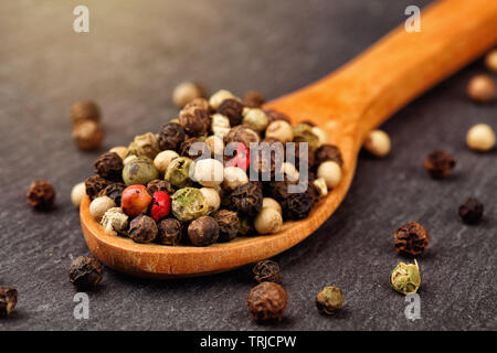 Macro shot of black, green, white and red peppercorn mix in a wooden spoon on a dark stone kitchen board with sun haze light Stock Photo