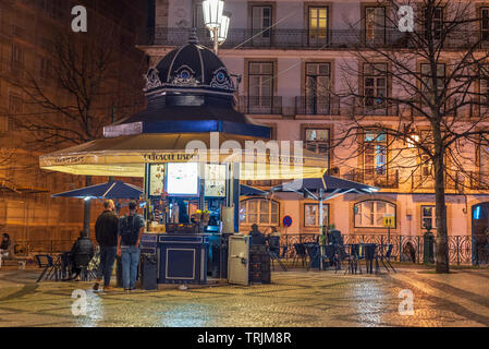 Two men buying at a kiosk located in Luis de Camoes square in downtown Lisbon, Portugal, at night, with traditional building backdrop. Stock Photo
