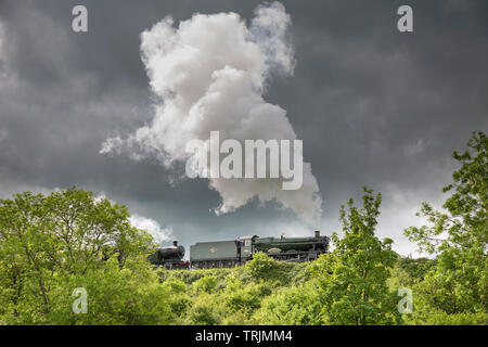 Side view of vintage UK steam train passing through English countryside puffing out steam on GWSR heritage line, sunny afternoon but dark clouds above. Stock Photo