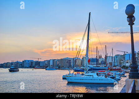 SLIEMA, MALTA - JUNE 19, 2018: The evening walk along the Sliema waterfront with a view on line of modern buildings, sail yachts and the bright sunset