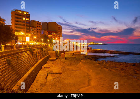 The evening walk along the seaside promenade of Sliema with a view on modern tourist neighborhoods, stone beach and twilight sky, Malta Stock Photo