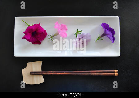 Four fresh bright petunia flowers on the white rectangular plate with wooden chopsticks on the black background Stock Photo