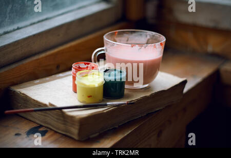 Still life of gouache paints, brushes, paint dilution cup and a book that teaches drawing. All items are on the old wooden windowsill. Stock Photo