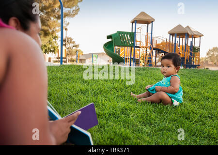 A woman holding a children's book tries to engage the attention of a baby girl sitting in the lawn of a kids playground. Stock Photo