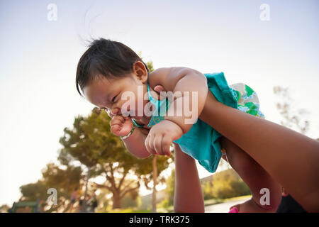 Baby girl being lifted up by mother into the sky, baby looks excited and nervous. Stock Photo