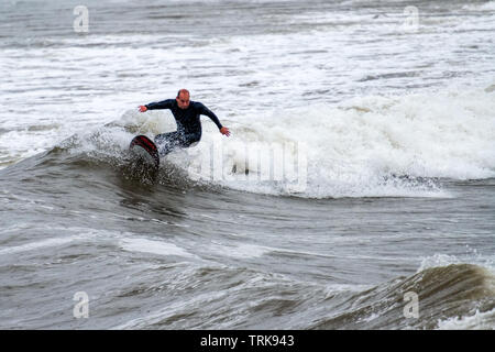 A middle-aged surfer riding the waves in the rain. Rest Bay, Porthcawl, UK. Stock Photo