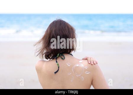 Outdoor summer portrait of Young Asian tan woman wearing bikini sitting on the beach applying sunscreen  with sun drawn on shoulder, summer vacation. Stock Photo