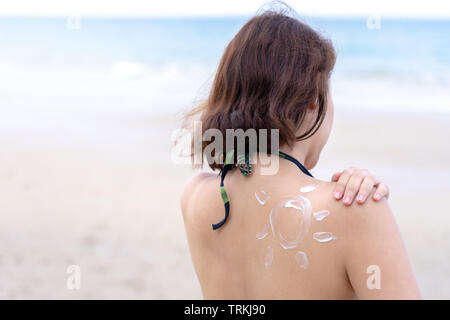 Outdoor summer portrait of Young Asian tan woman wearing bikini sitting on the beach applying sunscreen  with sun drawn on shoulder, summer vacation. Stock Photo