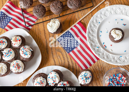 Overhead shot of mini cupcakes with white frosting and red, white, and blue sprinkles.  Additional cupcakes unfrosted on wire rack.  Wooden table.  US Stock Photo