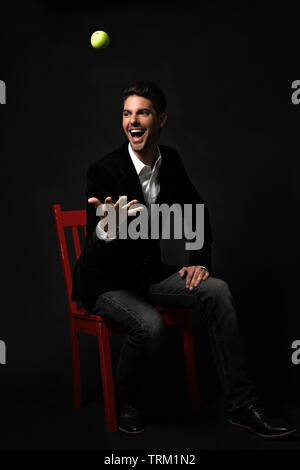 A good looking white male model in a black formal jacket, sitting on a red chair, throwing a green apple into the air, smiling and feeling excited! Stock Photo