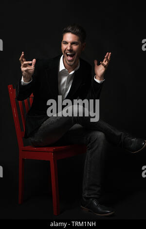 A good looking white male model modeling in a formal jacket, sitting on a red chair crossed legs. He throws his hands up in excitement and screaming! Stock Photo