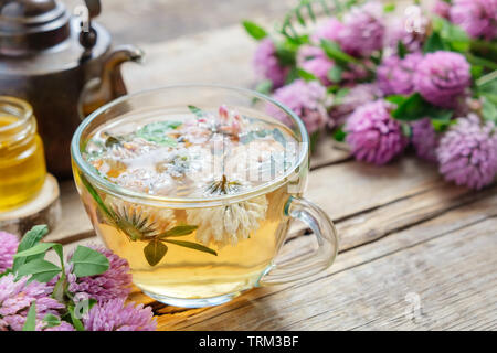 Red clover flowers, healthy herbal tea cup, honey jar and vintage copper tea kettle on table. Stock Photo