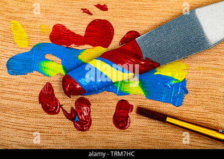 A mixed swirl of the primary colors red, yellow and blue, on a steel palette knife over a wooden palette with an artist's paint brush handy and ready Stock Photo