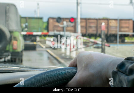 View of the driver hand on the steering wheel of the car, which stopped in front of a closed railway crossing at a red traffic light. The driver waits Stock Photo