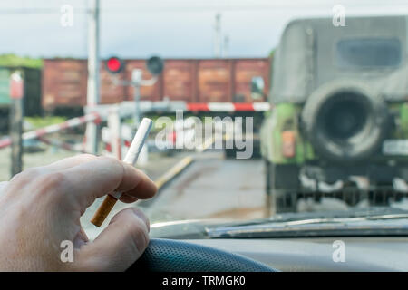 View of the driver hand with a cigarette on the steering wheel of the car, which stopped before a closed railway crossing at a red light Stock Photo