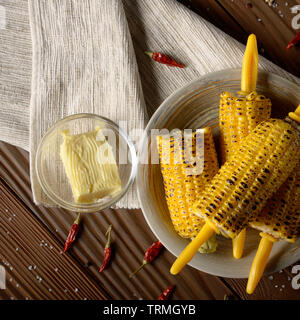 High angle view of Wooden table with deep grilled sweet corn cobs under melting butter with plastic holder on clay dish Stock Photo