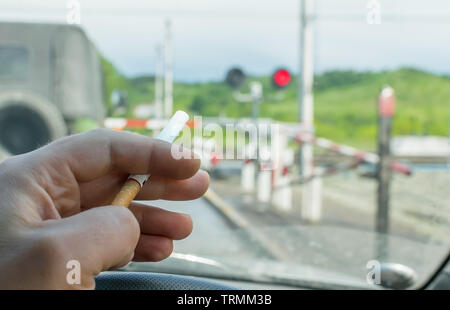 View of the driver hand with a cigarette on the steering wheel of the car, which stopped before a closed railway crossing at a red light Stock Photo