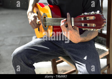 Young flamenco guitarist playing the guitar sitting in a chair in the street on a sunny day. Close up of a man playing the spanish guitar, chord in F. Stock Photo