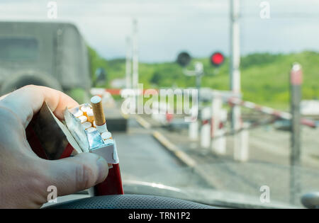 View of the driver hand with a pack of cigarettes on the steering wheel of the car, which stopped before a closed railway crossing at a red light Stock Photo
