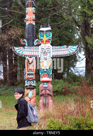 Stanley Park, Vancouver, British Columbia, Canada - Smiling Chinese tourist in foreground of colourful painted totem poles in woodland setting daytime Stock Photo