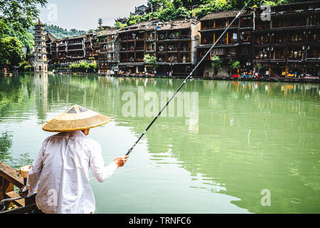 Chinese fisherman wearing an Asian conical hat and scenery of Fenghuang ancient phoenix town in background in Hunan China Stock Photo