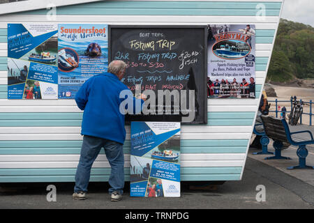Fisherman operating sea fishing trips writing the boat trip times on a blackboard by the seafront at Saundersfoot in Pembrokeshire, Wales Stock Photo