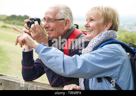 Senior Couple Hiking In Countryside Standing By Gate  And Taking Photo With Camera Stock Photo