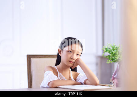 9 year-old little asian girl sitting at desk thinking with hands on cheek. Stock Photo