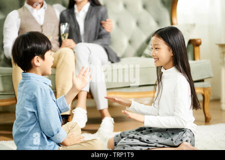 asian brother and sister sitting on carpet playing while parents watching in background Stock Photo