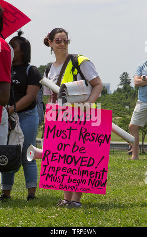 Washington, D.C., USA. 1st June, 2019. Woman wearing dangling earrings and reflective sunglasses, her head turned to the side, stands on green grass lawn carrying a lowered megaphone and a collection of signage, while other partially visible people appear around her, at the National March To Impeach on 1 June, 2019, on the Washington Monument Grounds in Washington, D.C., USA, organized by People Demand Action. The unfurled hot pink sign covering the lower half of her body reads, 'Trump must resign or be removed! Impeachment must begin now www.BAMN.com'. Kay Howell/Alamy Stock Photo
