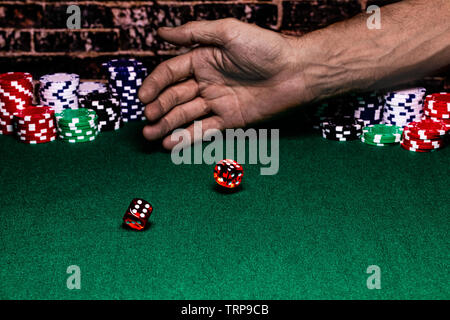 a close up of a pair of dice rolled by someone on a green felted gaming table.  Behind the throwing hand there are stacks of betting chips Stock Photo