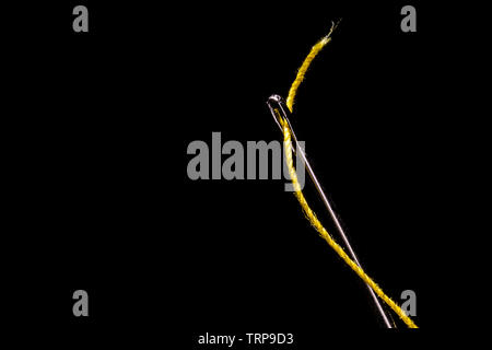 A macro image of a needle’s eye threaded with a piece of yellow thread on a field of solid black.  Focus is crisp on all elements of the shot and ther Stock Photo