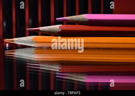 Orange, red, and pink color pencils on a background of black lead pencils. Stock Photo