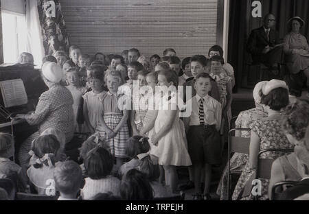 1955, historical, In a British village primary school, by a lady playing the piano, a group of young children singing a song in front of watching parents and other pupils, England, UK. Stock Photo