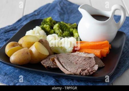 Lamb roast Sunday lunch dinner with vegetables on a aquare black plate, with gravy jug, on a blue tea towel.  White wood background Stock Photo