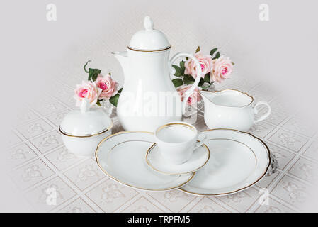 White crockery for coffee: coffeepot, cup, serving plate and sugar bowl as well as rose flowers are on a beautiful tablecloths Stock Photo