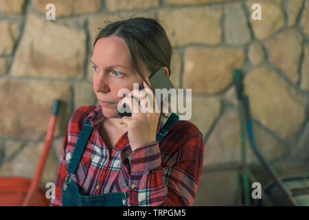 Worried female farmer talking on mobile phone in the stable. Adult woman in plaid shirt and denim overall trousers in telephone conversation. Stock Photo