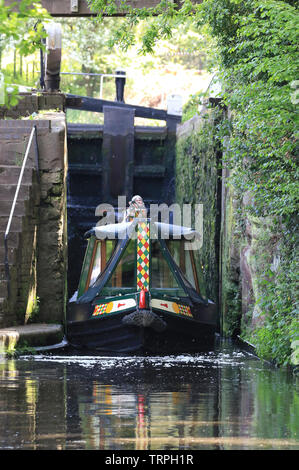 Portrait, front view of isolated UK narrow boat exiting canal lock on a sunny morning, steered by isolated female at rear of canal boat. Stock Photo