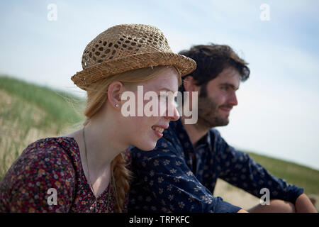 Beautiful young couple sitting on a hill on top of a dune enjoying the view and ocean breeze at the beach in bright summer sunshine Stock Photo