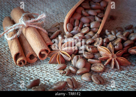 Roasted coffee beans spilling from a wooden scoop, cinnamon sticks and star anise spice on a on jute sack in a close up view Stock Photo