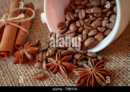Roasted coffee beans spilling from a cup, cinnamon with star anise spice on a on jute sack in a close up view Stock Photo