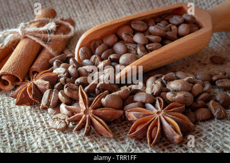 Roasted coffee beans spilling from a wooden scoop, cinnamon sticks and star anise spice on a on jute sack in a close up view Stock Photo
