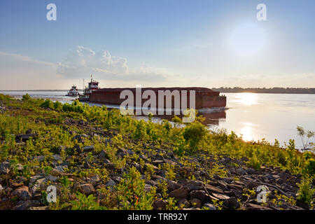 View from the riverbank of a loaded barge navigating the mighty Mississippi River with the help of a tug boat at sundown near Greenville, Mississippi Stock Photo