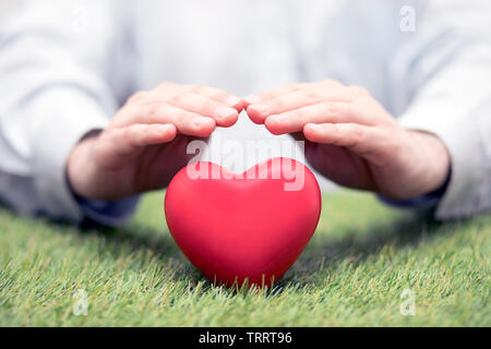 Red heart on green grass covered by hands. Health insurance or love concept Stock Photo
