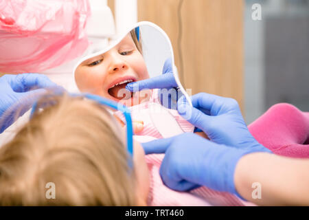 Dentist showing to little girl cured tooth in pediatric dental clinic. Child is sitting in a dental chair inspecting her teeth looking thru tooth-shap Stock Photo