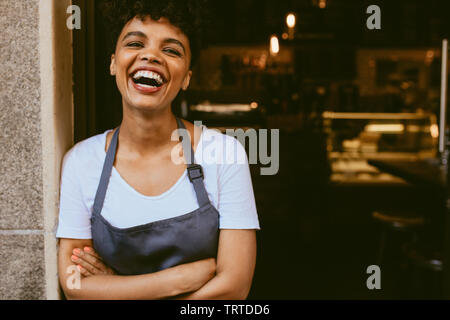 Cheerful female cafe owner standing at the door with her arms crossed. African woman in apron standing with her arms crossed and laughing. Stock Photo
