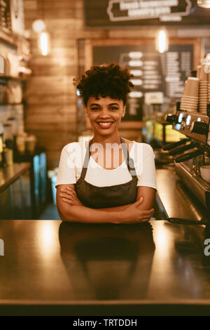 Smiling african woman in apron standing at cafe counter. Confident female barista standing behind counter. Stock Photo