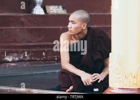 Yangon, Myanmar - March 2019: portrait of a young Buddhist male novice monk sitting at the porch in Shwedagon pagoda temple complex Stock Photo